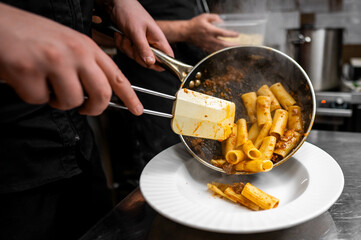 A chef serves freshly cooked rigatoni pasta onto a white plate, showcasing the vibrant sauce and ingredients. The kitchen environment highlights the art of Italian cuisine in action. © pavel siamionov