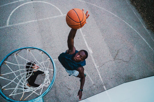 A basketball player attempts a dunk under the hoop on a concrete court, viewed from above, highlighting power, athleticism, and precision in an outdoor sports setting.