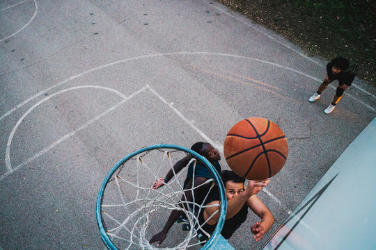 A vibrant capture of three basketball players engaging under the hoop on a marked outdoor court framed by bright trees and open sky.