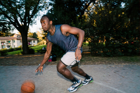 Black young man wearing athletic shorts and a tank top dribbles a basketball on an outdoor court surrounded by trees and nearby residential buildings during a sunny afternoon. flash light motion blur