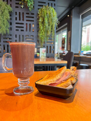 Hot chocolate in a clear glass cup and a warm ham and cheese sandwich served on a wooden table in the cafeteria. Blurred background.