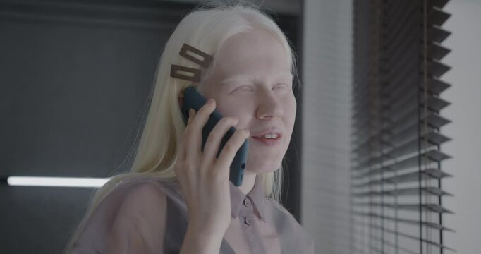 Cheerful albino woman talking on mobile phone smiling standing in stylish apartment alone. Wireless communication and people concept.