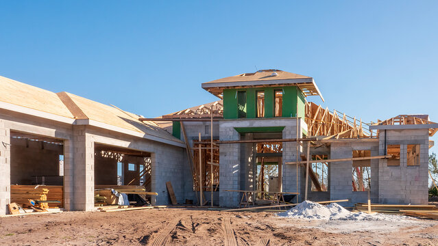 Street view of upscale single-family house under construction, with stacks of lumber in garages (left), and roof in progress, in a suburban residential development on a sunny day in southwest Florida