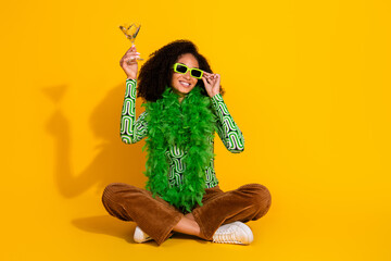 Young woman in vibrant green feather boa and shades holding cocktail on yellow background with playful, stylish vibe
