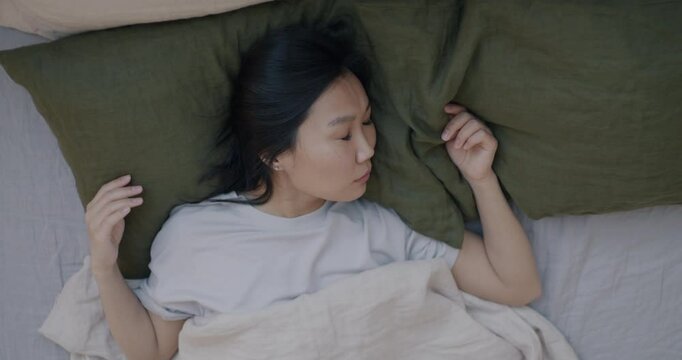 Top view of Asian woman sleeping in bed enjoying rest on pillow and comfortable sheets moving in sleep in bedroom at home. Rest and bedding concept.
