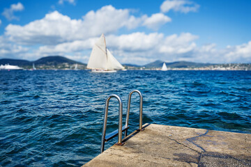 Handrail of a metal chrome ladder leading from a platform into the blue sea in the background sails a historic yacht.