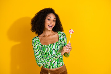 Joyful young woman with colorful lollipop against vibrant yellow backdrop, wearing a stylish green patterned top and smiling happily.