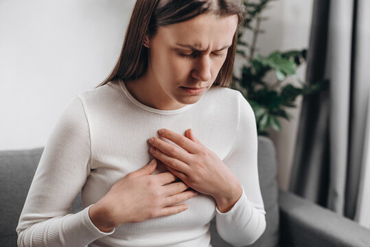 Close up of unhealthy young caucasian woman having severe chest pain while sitting alone on couch, sad worried female suffers from heartache at home, infarction or heart disease concept