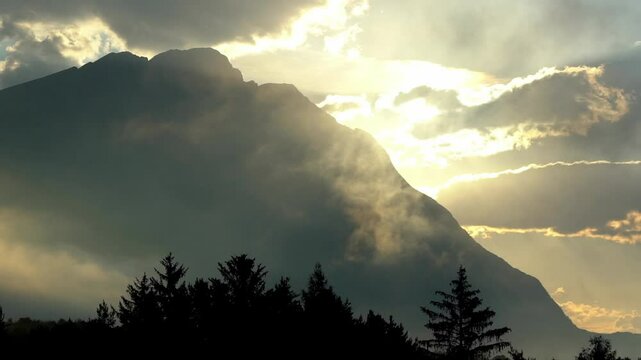 Foggy sunrise in the mountains with the sun peeking through rising mist and wandering clouds, creating a golden shimmer and an otherworldly atmosphere