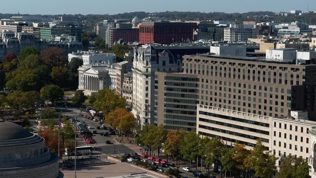 A aerial view of Washington DC cityscape with White House, US Treasury, government buildings, trees, and cars. The view includes a street, and multiple trees with fall foliage.