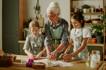 Senior woman baking with two children in kitchen, wearing matching aprons and making cookies on wooden countertop