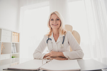 Smiling senior female doctor sitting confidently in her clinic office with a stethoscope, ready for the next medical consultation