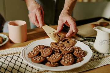 Hands arranging freshly baked chocolate chip cookies on a white plate in a cozy home kitchen setting, with other baking items around on a patterned tablecloth