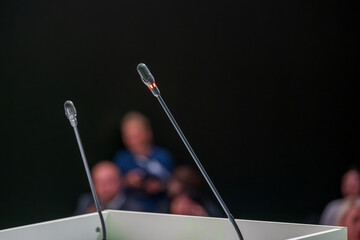 Close-up of microphones in focus at speaking event, audience blurred in background. Ideal for presentations, conferences, or discussions.