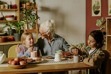 Senior woman with grandchildren sharing warm conversation and enjoying snacks around table. Cozy kitchen setting with plants and various household items enhancing ambiance