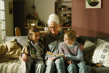 Elderly woman reading story to two granddaughters on couch in cozy living room illuminated by warm...