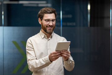 Smiling young male businessman standing in office wearing glasses and tie, holding and using tablet