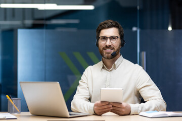 Portrait of a young businessman in a headset sitting at a workplace with a laptop, holding a tablet in his hands and looking at the camera with a smile