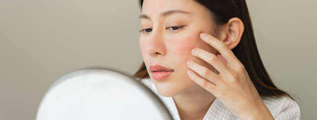 Asian young woman looking face skin in the mirror have a red rash on her face from cosmetic allergy