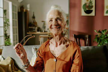 Elderly woman standing in living room, smiling while holding glasses. Cozy interior features...