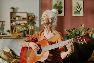 Senior woman smiling while playing acoustic guitar in cozy living room with plants and paintings in...