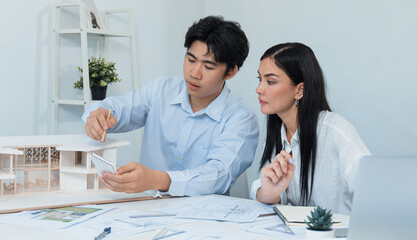 Professional male asian architect using ruler to measure house model length while young beautiful caucasian colleague using laptop to analyzed data on meeting table with house model. Immaculate.