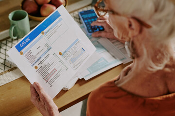 Obraz premium Senior woman examining gas bill and calculating expenses while sitting at dining table. Home setting includes papers, calculator, fruit bowl, and green cup