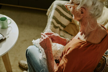 Senior woman sitting indoors while crocheting with focused expression. Warm lighting providing cozy...