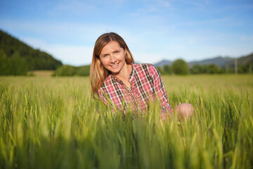 Young woman in gingham checkered shirt and shorts crouching in unripe green wheat field, smiling, blue sky and blurred landscape with small forest background