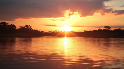 Fototapeta premium Vibrant hues of orange, pink, and yellow fill the sky as the sun sets over a tranquil lake. Gentle ripples reflect this breathtaking display, creating a peaceful ambiance.