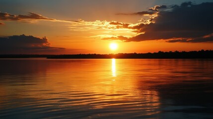 Fototapeta premium Vibrant hues of orange, pink, and yellow fill the sky as the sun sets over a tranquil lake. Gentle ripples reflect this breathtaking display, creating a peaceful ambiance.