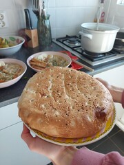 A freshly baked pita bread, still warm and soft, being held up by a hand. The bread is round and has a golden-brown crust with sesame seeds. In the background, there are bowls of soup and other dishes