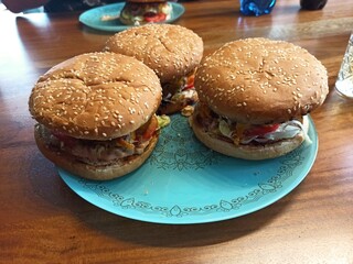 Three homemade hamburgers sit on a decorative blue plate. Each burger features a sesame seed bun, grilled meat, and an assortment of fresh vegetables, embodying a delightful meal experience