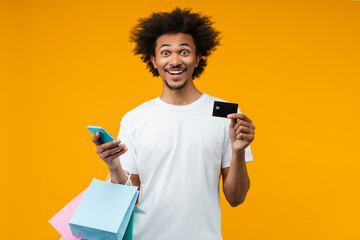 Portrait of joyful young man customer posing in studio with a bunch of shopping bags, credit card and smartphone in hands with amazed face expression, isolated over orange yellow background