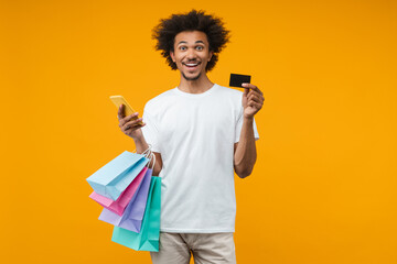 Excited young man customer posing in studio over orange yellow background with a bunch of shopping bags, credit card and smartphone in hands