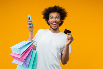 Joyful happy smiling young man customer posing in studio with a bunch of shooping bags, credit card and mobile phone in hands