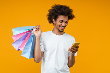 Studio portrait of handsome smiling African American young man with shopping bags in hand using his smart phone over bright yellow background