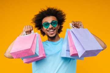 Close up portrait of joyful young man customer in trendy heart shaped sunglasses posing in studio with bunches of shopping bags in hands isolated over bright colored orange yellow background