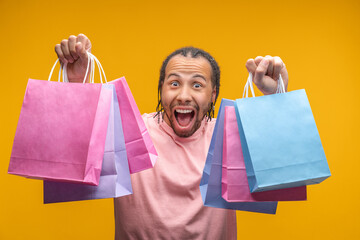 Studio portrait of euphoric young man customer posing with bunches of shopping bags in his hands with amazed open mouth face expression, isolated over yellow background