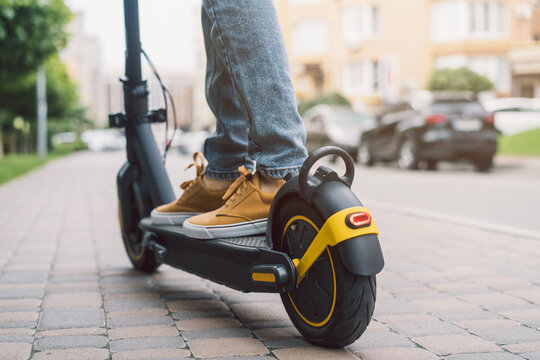 Close up cropped low angle photo of the feet of unrecognizable man standing by or riding electric kick scooter e-scooter outdoors with the street on the background