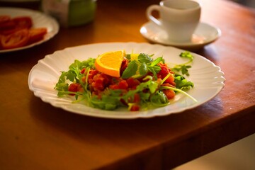 White plate with tuna tartare, a touch of arugula, orange, lime, and mint on a wooden table, surrounded by breakfast dishes.