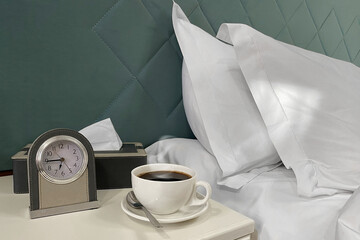 - A cup of hot coffee, a clock, and a box of tissues stand on a nightstand near the bed with white pillows.