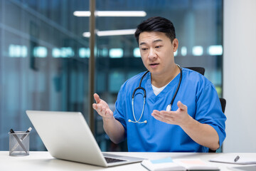 Asian man doctor wearing blue scrubs engaged in video call on laptop, illustrating remote healthcare communication. Gestures convey professionalism during online telemedicine consultation.