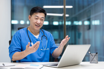 Asian man doctor conducting virtual consultation with laptop. Stethoscope and blue scrubs indicate healthcare professional engaging in online patient communication.