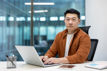 Asian man businessman using laptop at office desk, looking confident. Surroundings include smartphone and notes. Modern workspace evokes professionalism, focus, determination, productivity.