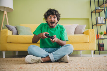 Full length portrait of nice young man sit carpet floor hold controller play game wear t-shirt modern green interior flat indoors © deagreez