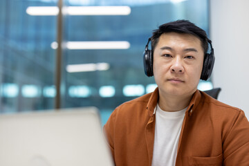 Asian man businessman wearing headphones in modern office environment focused on work. Seated at...