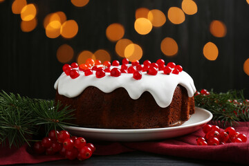 Tasty Christmas cake with red currants and fir branches on black wooden table against blurred lights, closeup