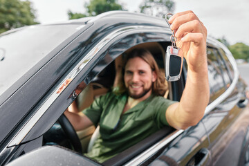 Young man holding car keys inside a vehicle with a joyful expression and greenery in the background