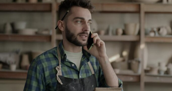 Pottery studio manager talking to client on mobile phone then writing business information in notebook. Young man in apron developing workshop.